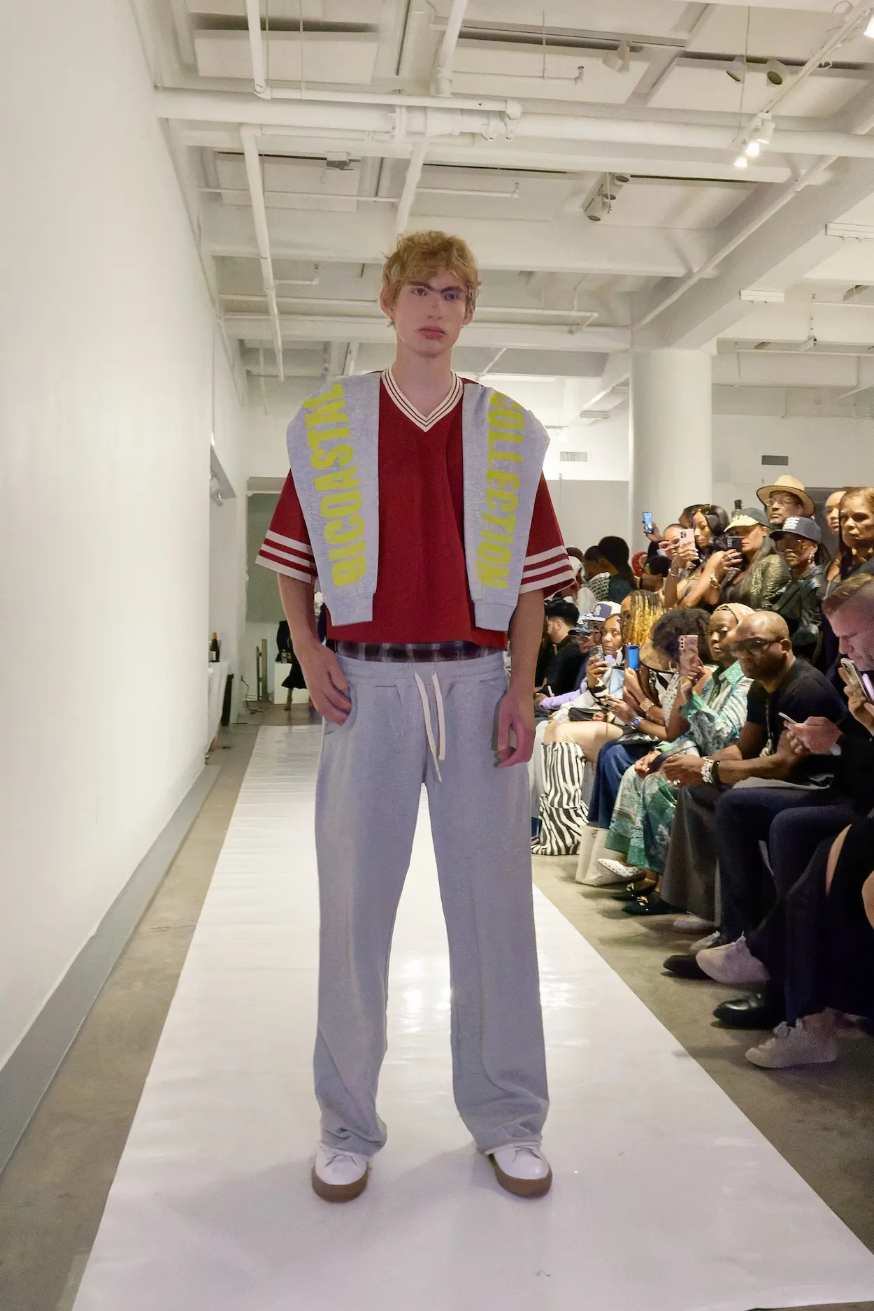 A model wears a red sports-inspired top, gray wide-leg trousers, and a BICOASTAL COLLECTION graphic layer during The Impossible Dream SS27 show.