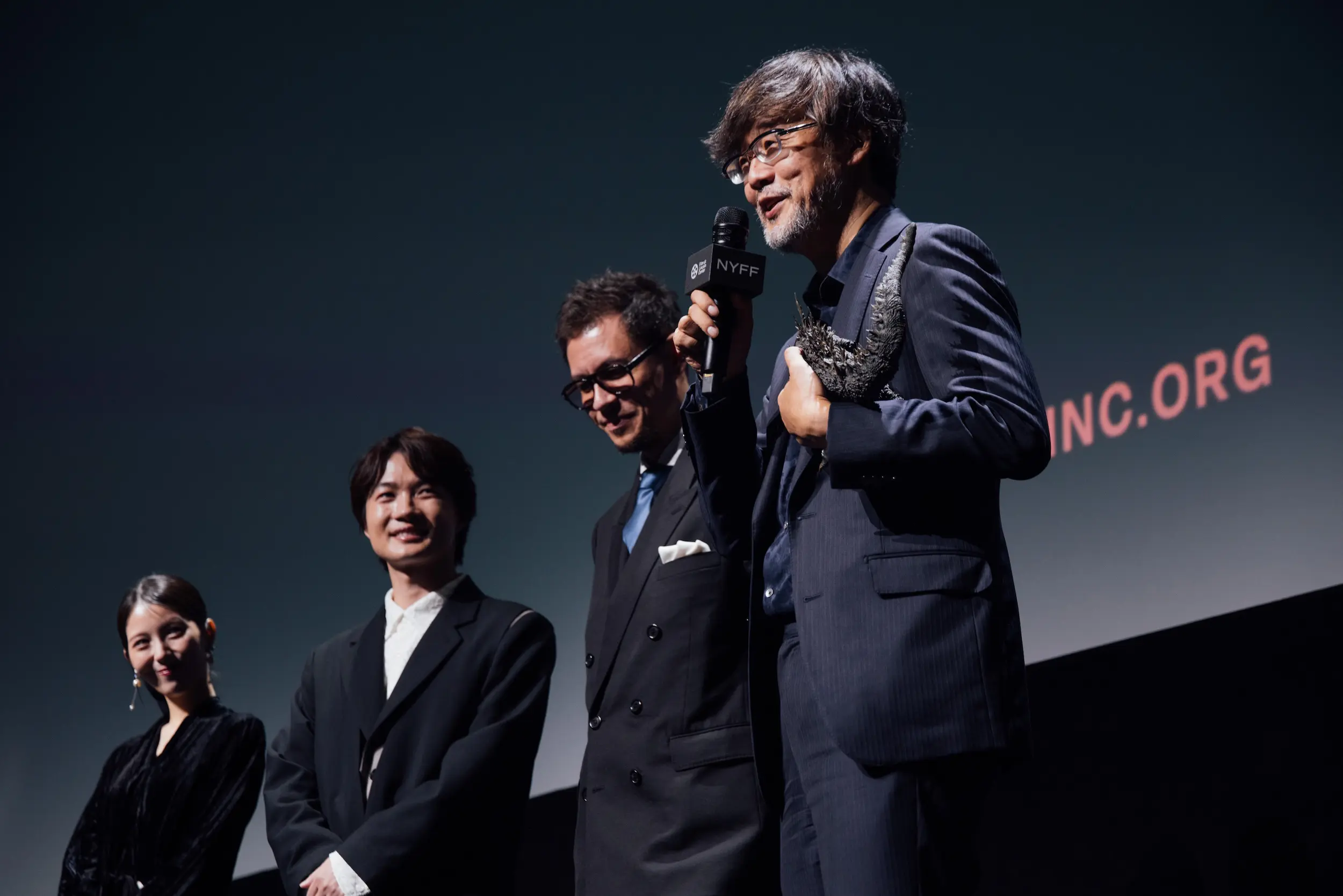 Takashi Yamazaki speaks onstage while Ryunosuke Kamiki and Minami Hamabe stand nearby before the Godzilla Minus Zero premiere screening at NYFF64.