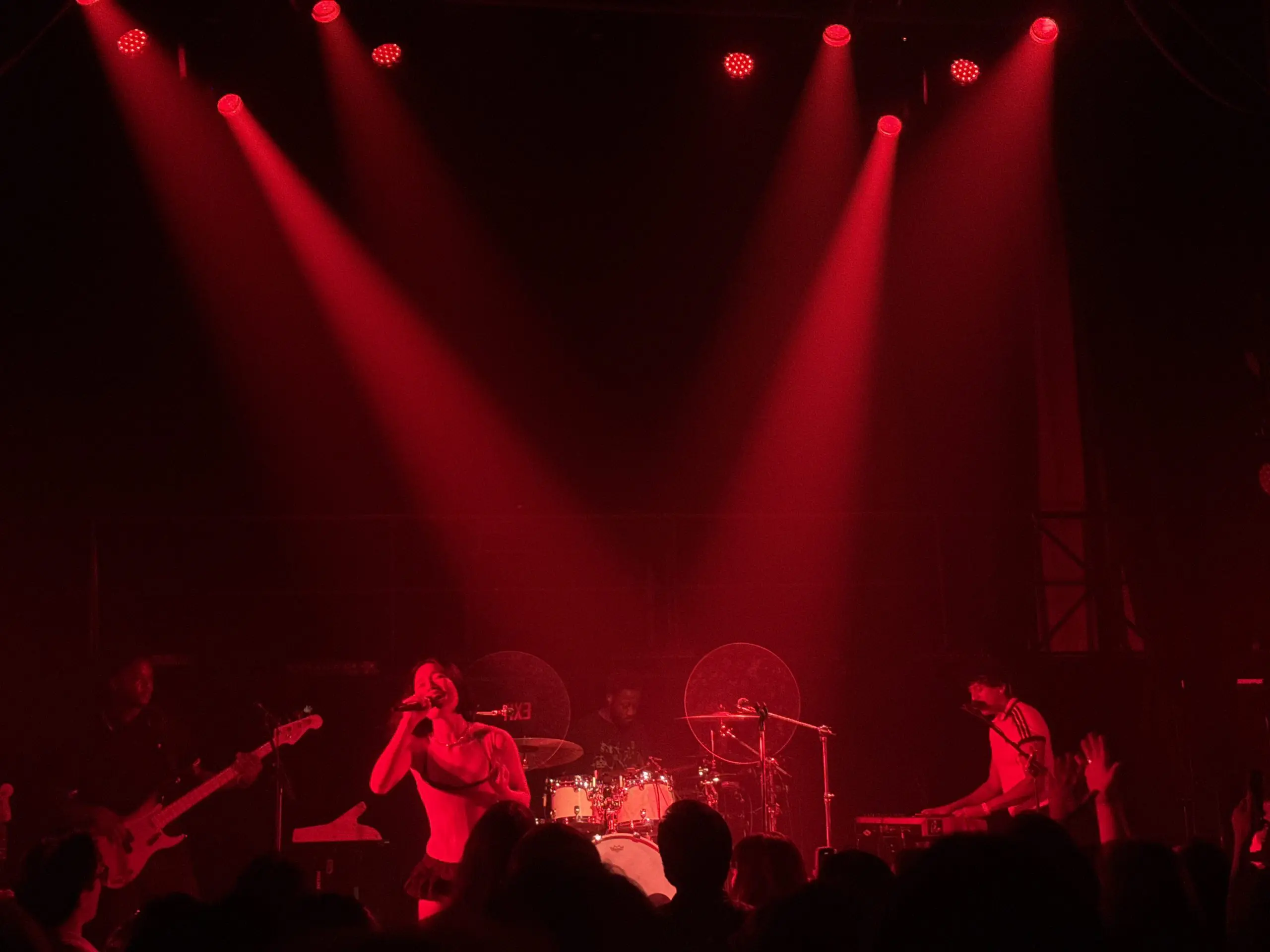 Chloe Qisha singing into a microphone in red stage light, with a bassist, drummer, and keyboardist on stage.
