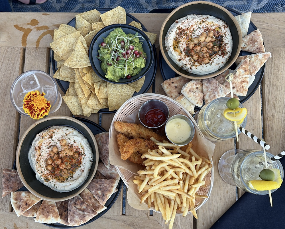 Selection of appetizers served during Lynette Marrero's cocktail menu tasting at The Roof at PUBLIC New York, featuring hummus, guacamole, pita chips, crispy chicken tenders, and fries.